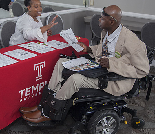 image of a man and a woman sitting at a table at a job fair. The man is recievieng information about working for Temple University. He is in a wheelchair.